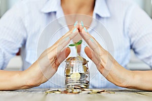 BusinesswomanÃ¢â¬â¢s palms protecting a green plant growing from a jar full of cash money, savings or incomes protection concept